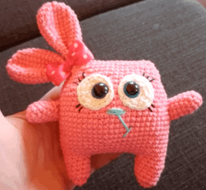 Roztomilá pletená hračka s velkýma očima a růžovým motivem, ideální jako dárek nebo dekorace.