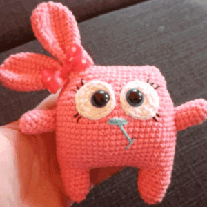 Roztomilá pletená hračka s velkýma očima a růžovým motivem, ideální jako dárek nebo dekorace.